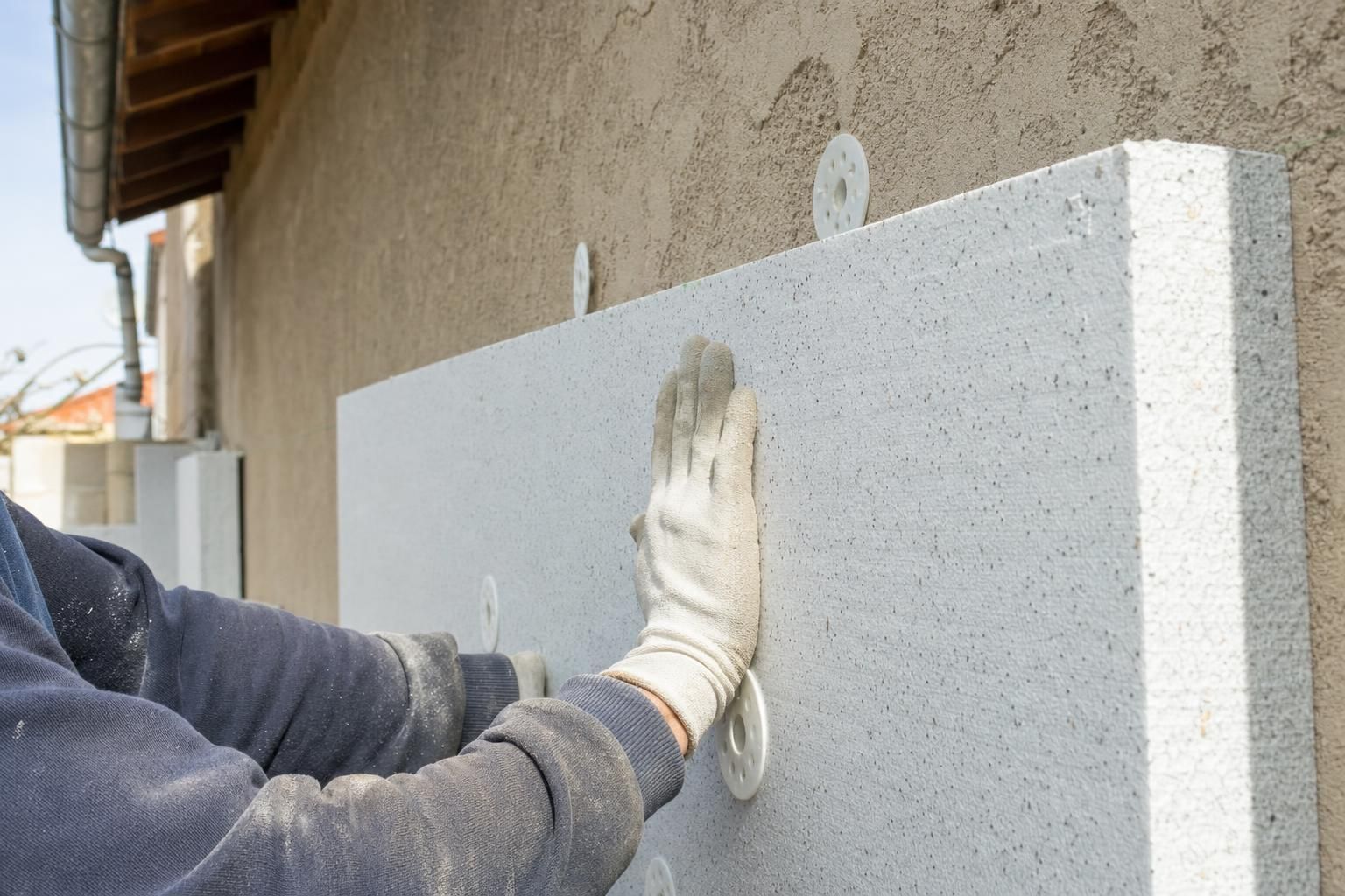 Mains gantées d'un artisan posant un panneau d'isolation thermique extérieure sur la façade d'une maison individuelle, lumière naturelle.
