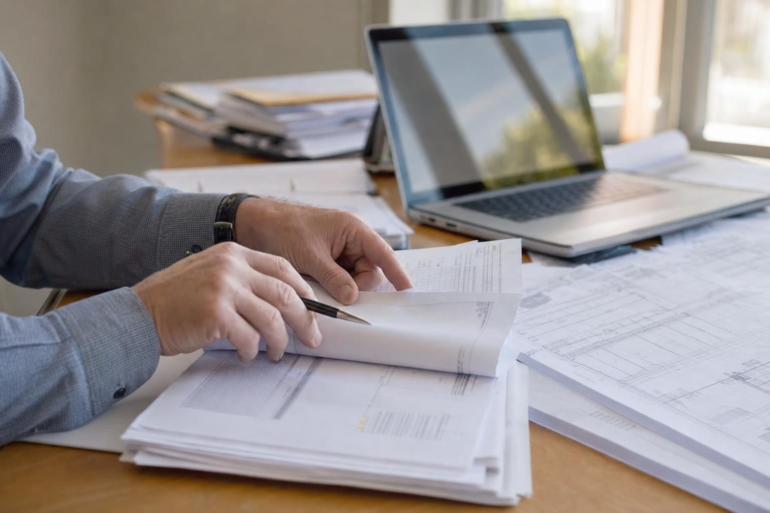 Gros plan sur des mains feuilletant des documents contractuels et devis sur un bureau de PME, ordinateur ouvert et plans de chantier en arrière-plan, lumière naturelle.