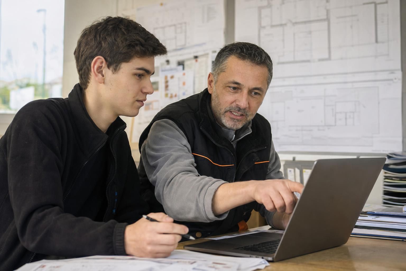 Artisan expérimenté montrant à un jeune collègue une procédure sur un ordinateur portable dans un bureau de PME du bâtiment, plans au mur en arrière-plan, lumière naturelle.
